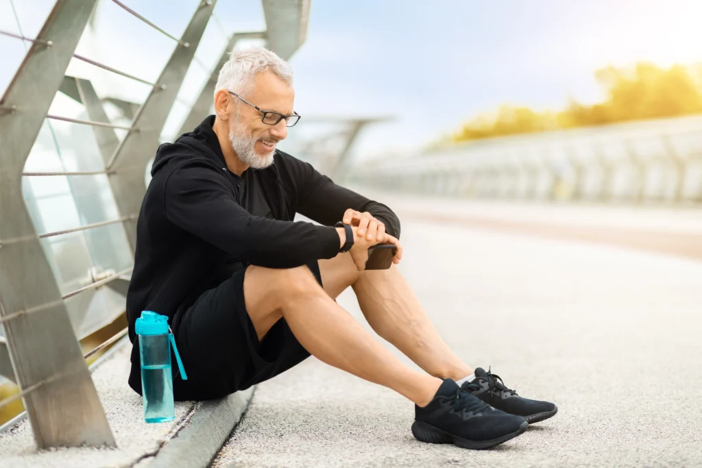 attraktiver Sportler sitzt auf dem Boden an einer Brücke, überprüft seine Smartwatch, trinkt Wasser während des Outdoor-Workouts und verfolgt seine Trainingsergebnisse, Freiraum