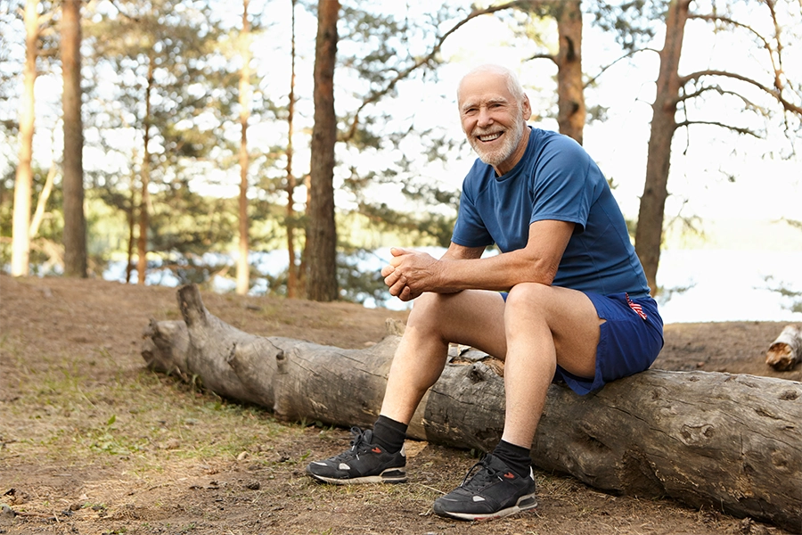 Horizontale Aufnahme glücklicher fröhlicher älterer Rentner mit dichtem weißem Bart sitzt auf umgefallenem Baum im Wald, lacht freudig, macht Pause nach intensivem morgendlichem Cardio-Workout, trägt Turnschuhe