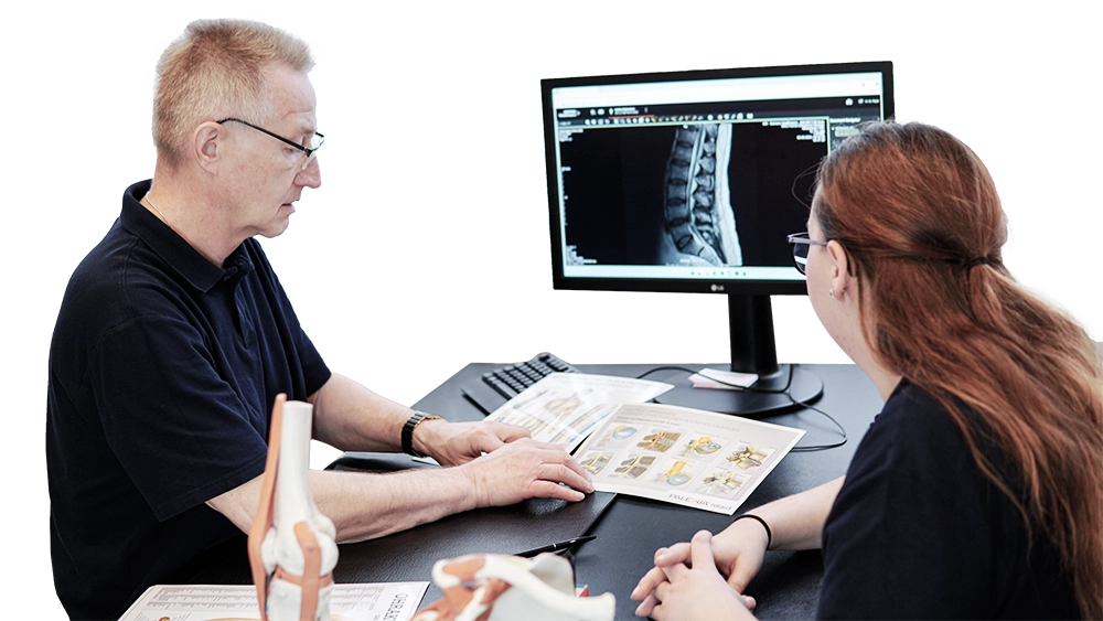 Dr. Pape erklärt einer Patientin anhand von Bildern der Wirbelsäule. Im Hintergrund sieht man einen Monitor mit einer Röntgenaufnahme.
