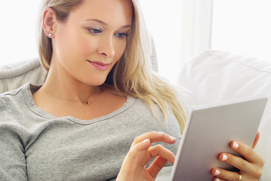 Frau lächelnd mit Tablet auf dem Sofa – entspannt, Streaming-Medien, Entertainment-App am Wochenende; weibliche Person, Abo-Plattform, Technologie zu Hause, E-Book, Website, Komfort im Wohnzimmer